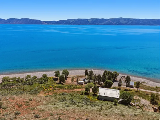 Waterfront property featuring a structure with a light-colored roof, surrounded by mature trees and natural landscaping, with a gravel beach at the water's edge and distant mountains