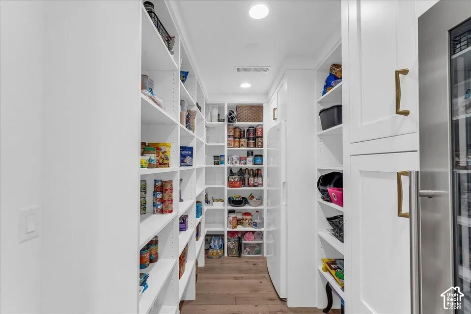 Spacious pantry featuring extensive built-in shelving, recessed lighting, wood-finish flooring, a stainless steel beverage refrigerator, and white cabinetry with brass hardware