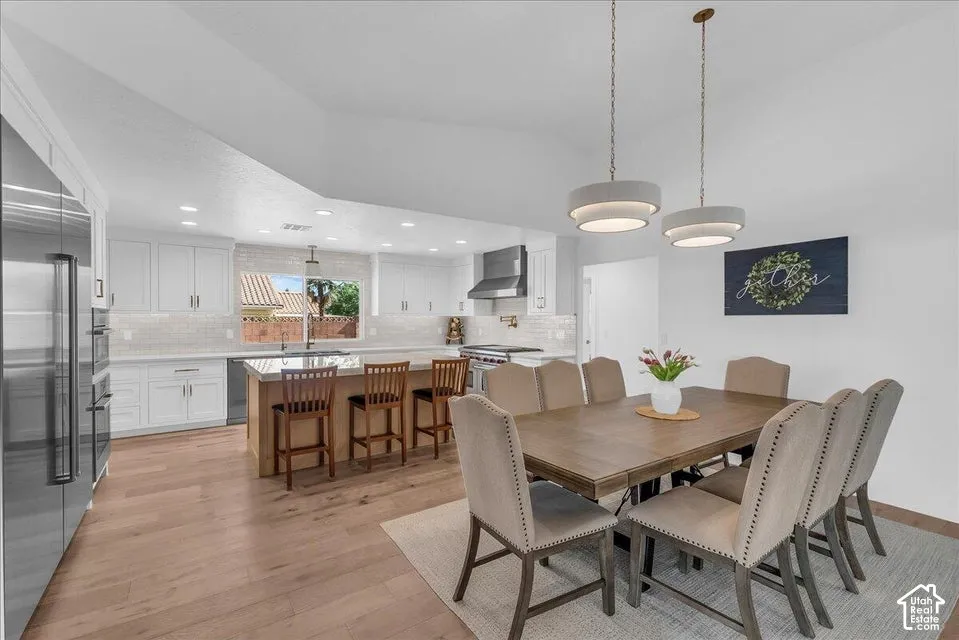 Spacious kitchen and dining area featuring light wood-finish flooring, white cabinetry, a large kitchen island with seating, a stainless steel refrigerator, and dual pendant lighting fixtures