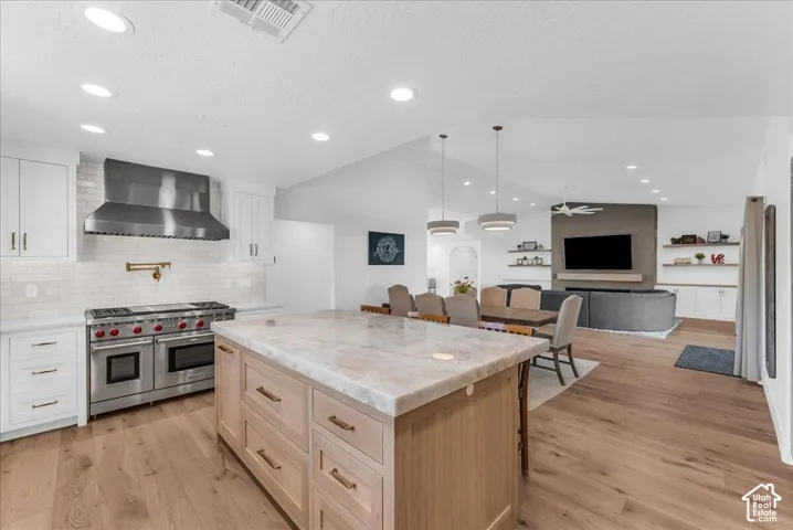 Spacious kitchen featuring a large central island with a stone countertop, light wood cabinetry with gold-tone hardware, and a stainless steel range with a professional-grade hood
