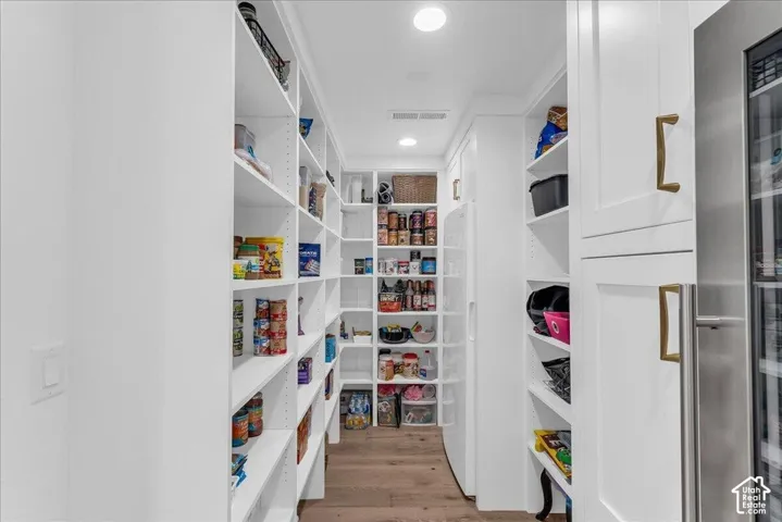 Spacious pantry featuring extensive built-in shelving, recessed lighting, wood-finish flooring, a stainless steel beverage refrigerator, and white cabinetry with brass hardware