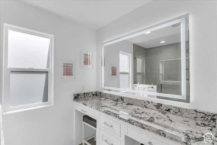 Vanity area featuring a large illuminated mirror, expansive stone countertop, white cabinetry with silver hardware, and a built-in makeup vanity