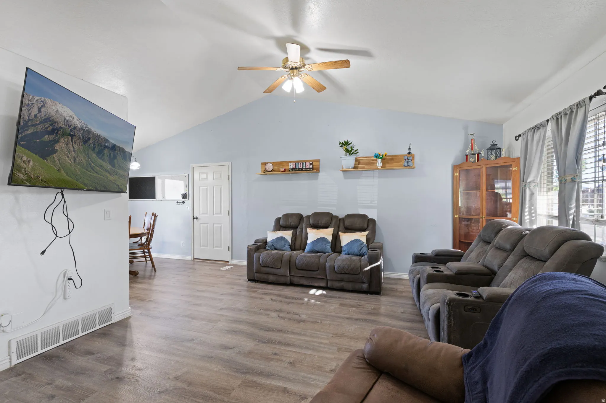 Vaulted ceiling with a ceiling fan, wood-finish flooring, light blue wall paint, window with blinds and curtains, and a white interior door