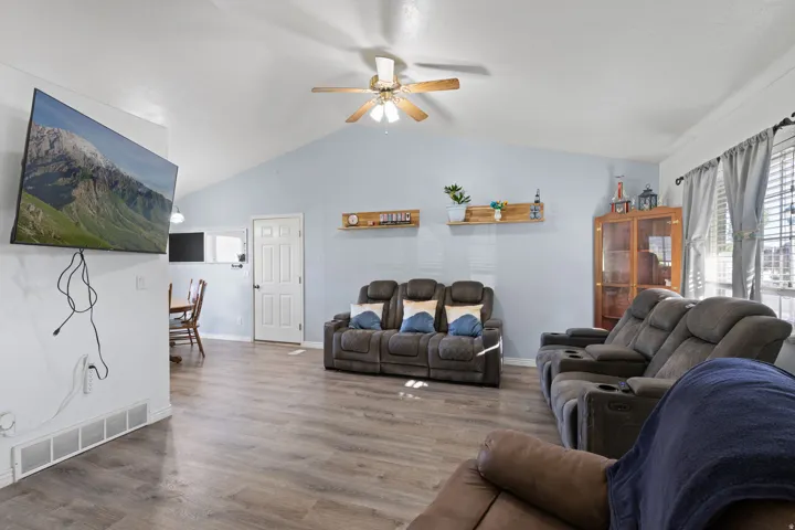 Vaulted ceiling with a ceiling fan, wood-finish flooring, light blue wall paint, window with blinds and curtains, and a white interior door