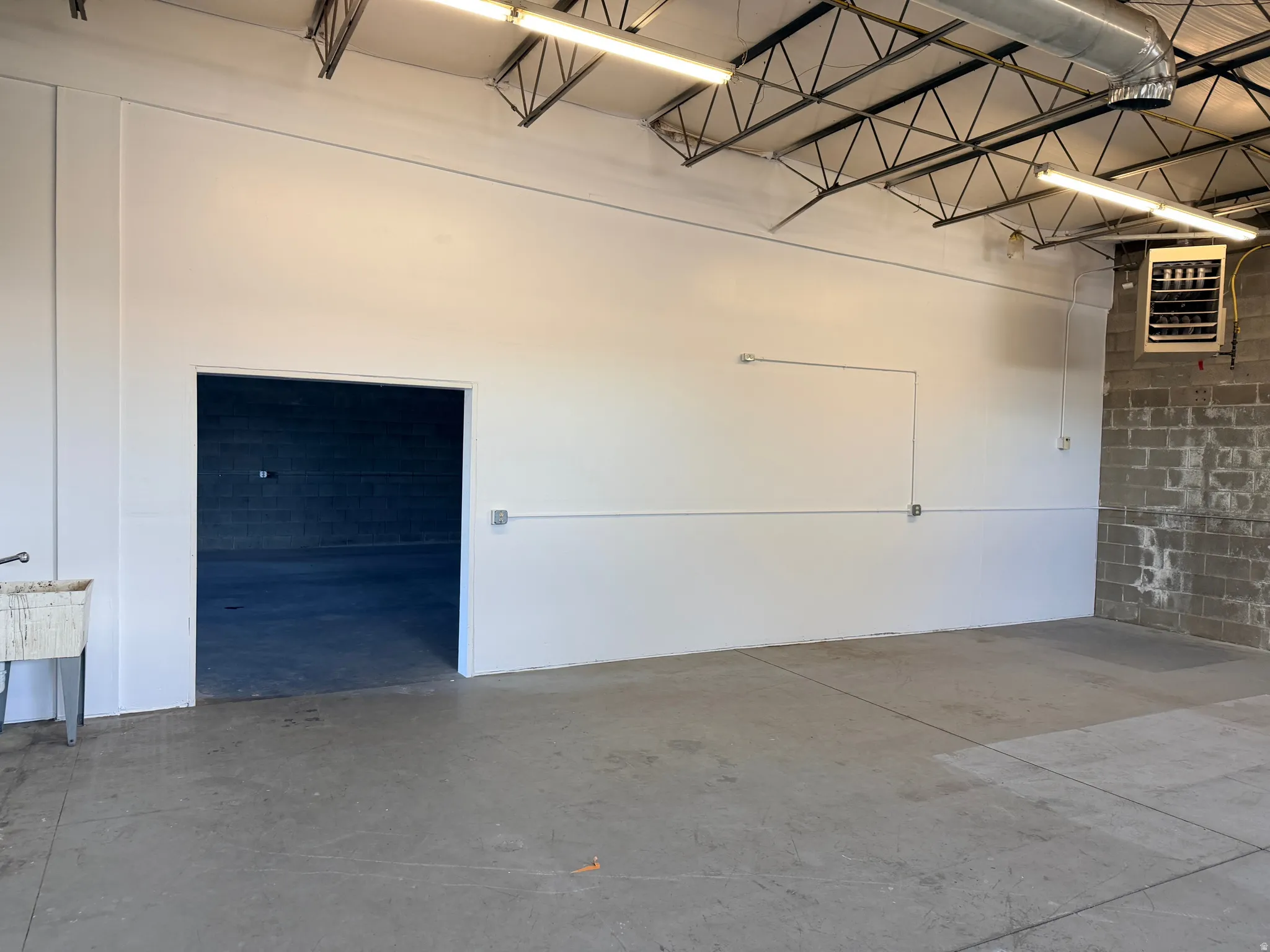 Interior commercial space featuring exposed ceiling trusses, concrete flooring, fluorescent overhead lighting, and a utility sink