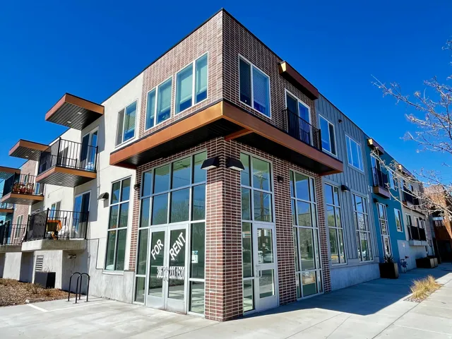 Modern commercial storefront featuring large glass windows, brick and panel siding, upper-level balconies, and a clear blue sky background