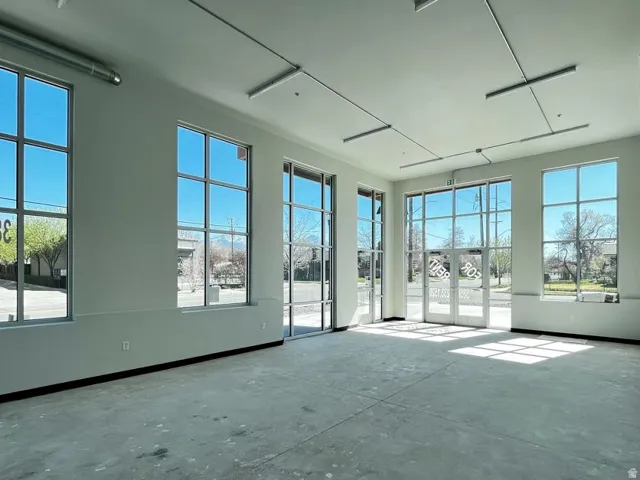Expansive commercial space featuring a concrete floor, multiple large windows with grid patterns, double glass entry doors, exposed ceiling ductwork, and track lighting fixtures
