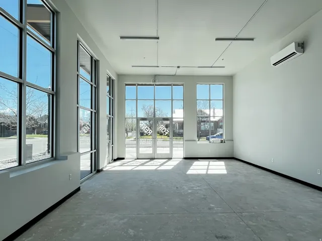 Open commercial space featuring multiple large windows and glass entry doors, high ceilings, concrete flooring, and wall-mounted climate control unit