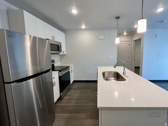 Kitchen featuring an island with an undermount sink and gooseneck faucet, stainless steel appliances, white cabinetry with silver hardware, recessed lighting, and wood-finish flooring