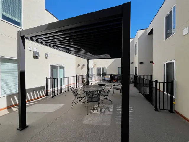 Spacious outdoor patio featuring a black louvered pergola, textured concrete flooring, black metal railing, and light-colored stucco exterior