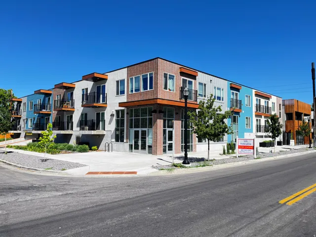 Contemporary multi-story building featuring varied panel siding, individual balconies with black railings, large ground-floor windows, an integrated concrete sidewalk, and street-level landscaping