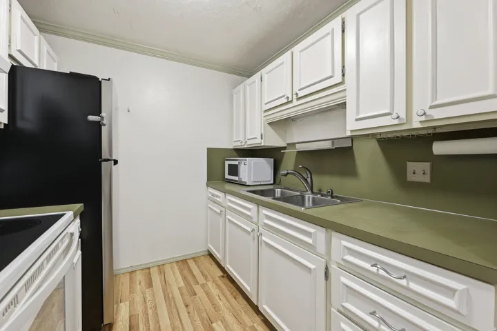 Compact kitchen with wood-finish flooring, white cabinetry, a double basin sink, solid surface countertops, and a black refrigerator