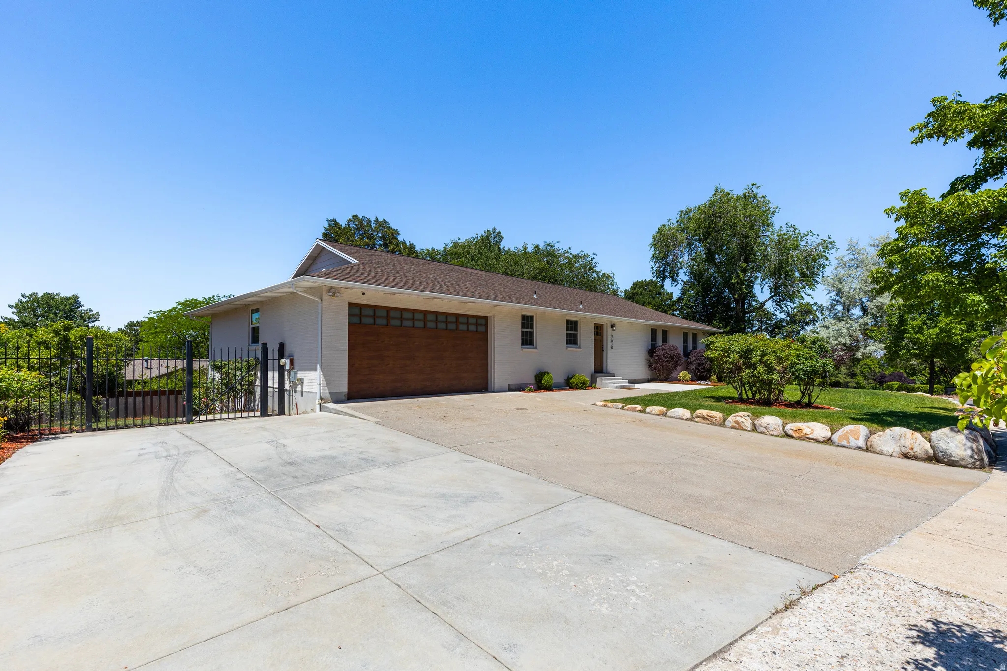 Stucco exterior with a wood-finish garage door, expansive paved driveway, wrought iron gate, and a landscaped lawn featuring natural stone edging