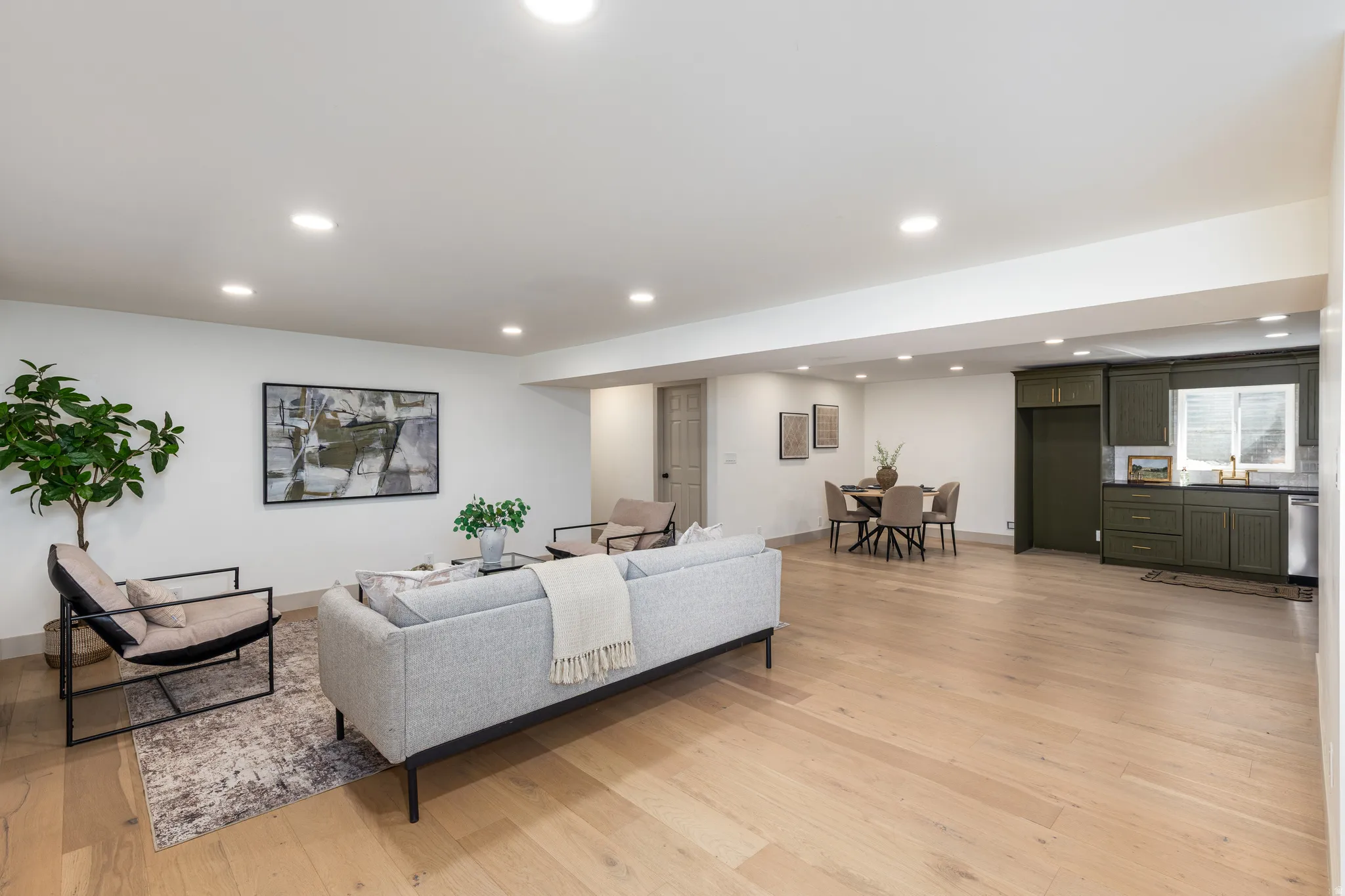 Open-concept living area featuring light wood-finish flooring, recessed lighting, white walls, a dining area, and a kitchen with dark green cabinetry and a window above the sink