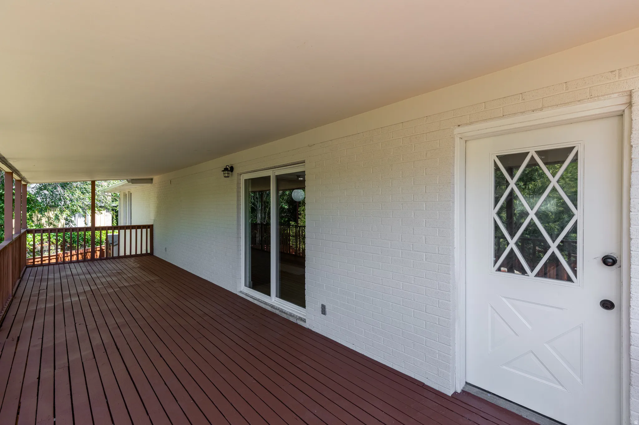 Expansive covered patio featuring wood-finish decking, painted brick exterior, and a white entry door with diamond-patterned lites