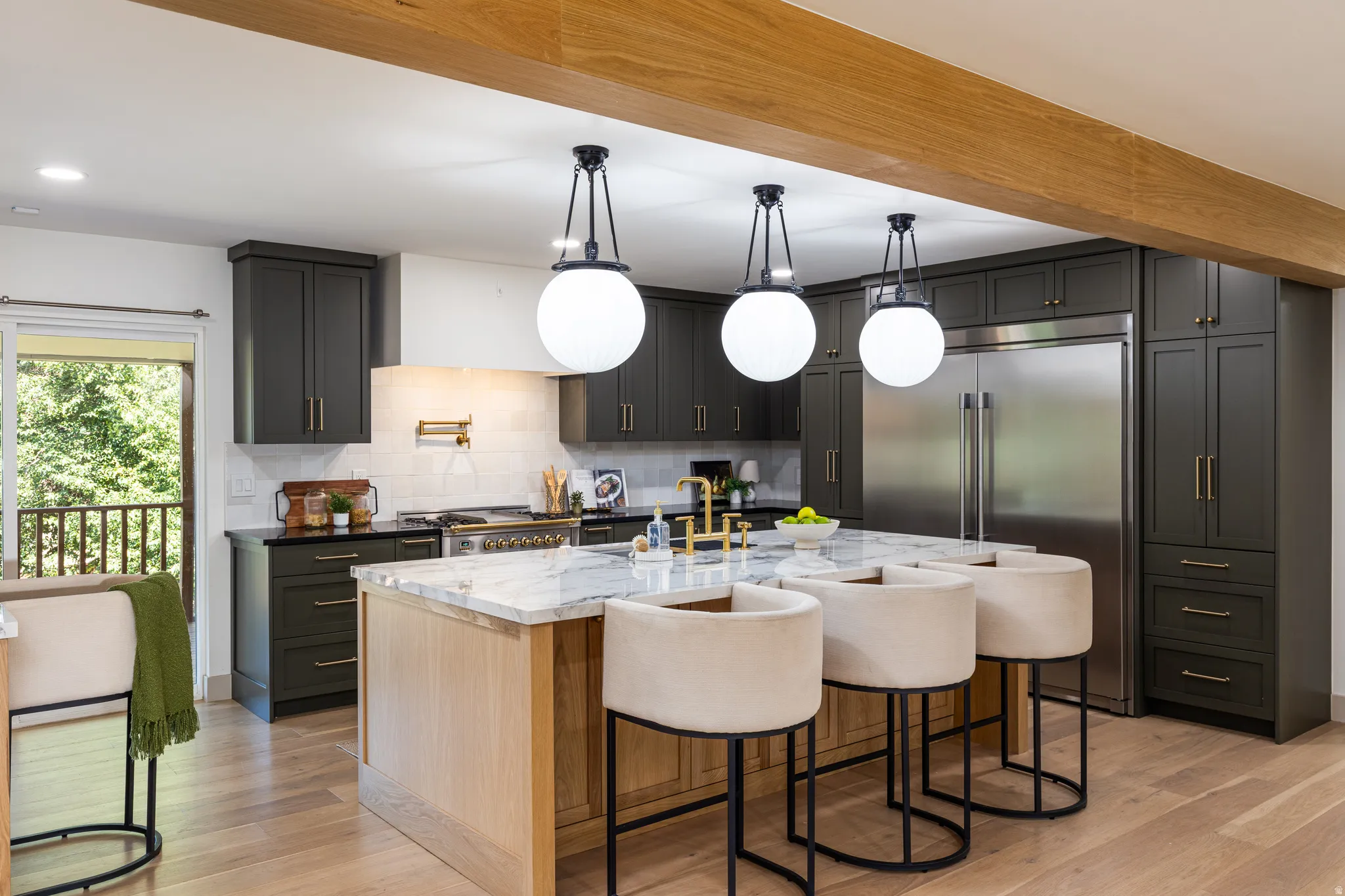 Contemporary kitchen featuring dark cabinetry, light wood-finish flooring, a large central island with seating, a stainless steel refrigerator, and a tiled backsplash with a pot filler