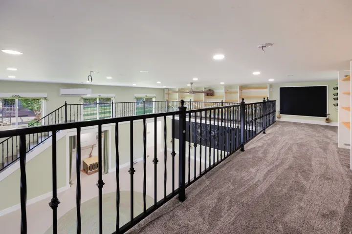 Spacious loft area featuring patterned carpeting, recessed lighting, built-in shelving, decorative iron railings, and an integrated projector screen