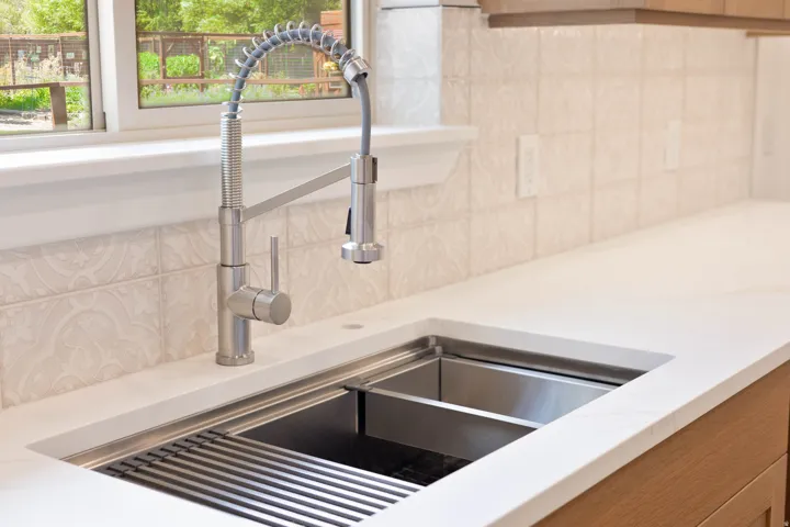 Stainless steel workstation sink with an integrated drying rack, white solid surface countertop, brushed nickel pre-rinse faucet, and decorative tile backsplash