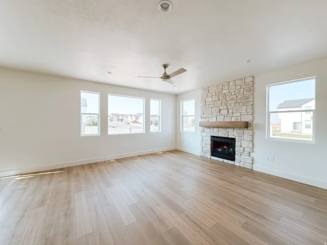 Spacious living area featuring light wood-finish flooring, a stone-clad fireplace with a wood mantle, numerous windows, and a ceiling fan