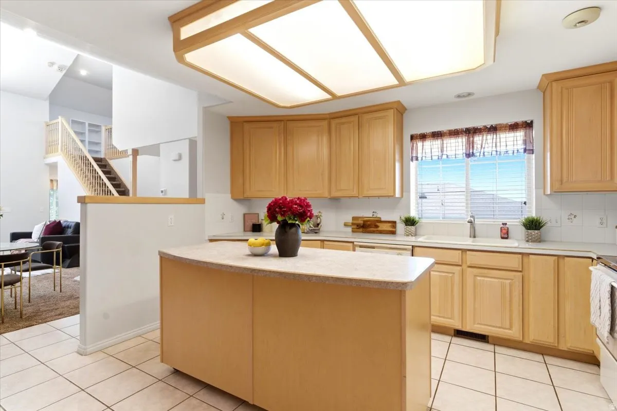 Kitchen featuring a moveable central island with a solid surface countertop, light wood-finish cabinetry, tile flooring, and a large window above the sink