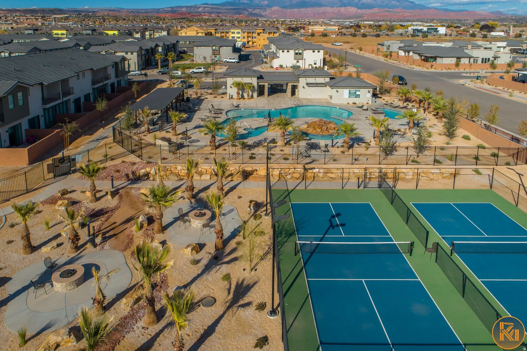 Community amenity area featuring two blue pickleball courts, a large swimming pool with rock features, a shaded lounging area, and multiple fire pit gathering spaces surrounded by desert landscaping