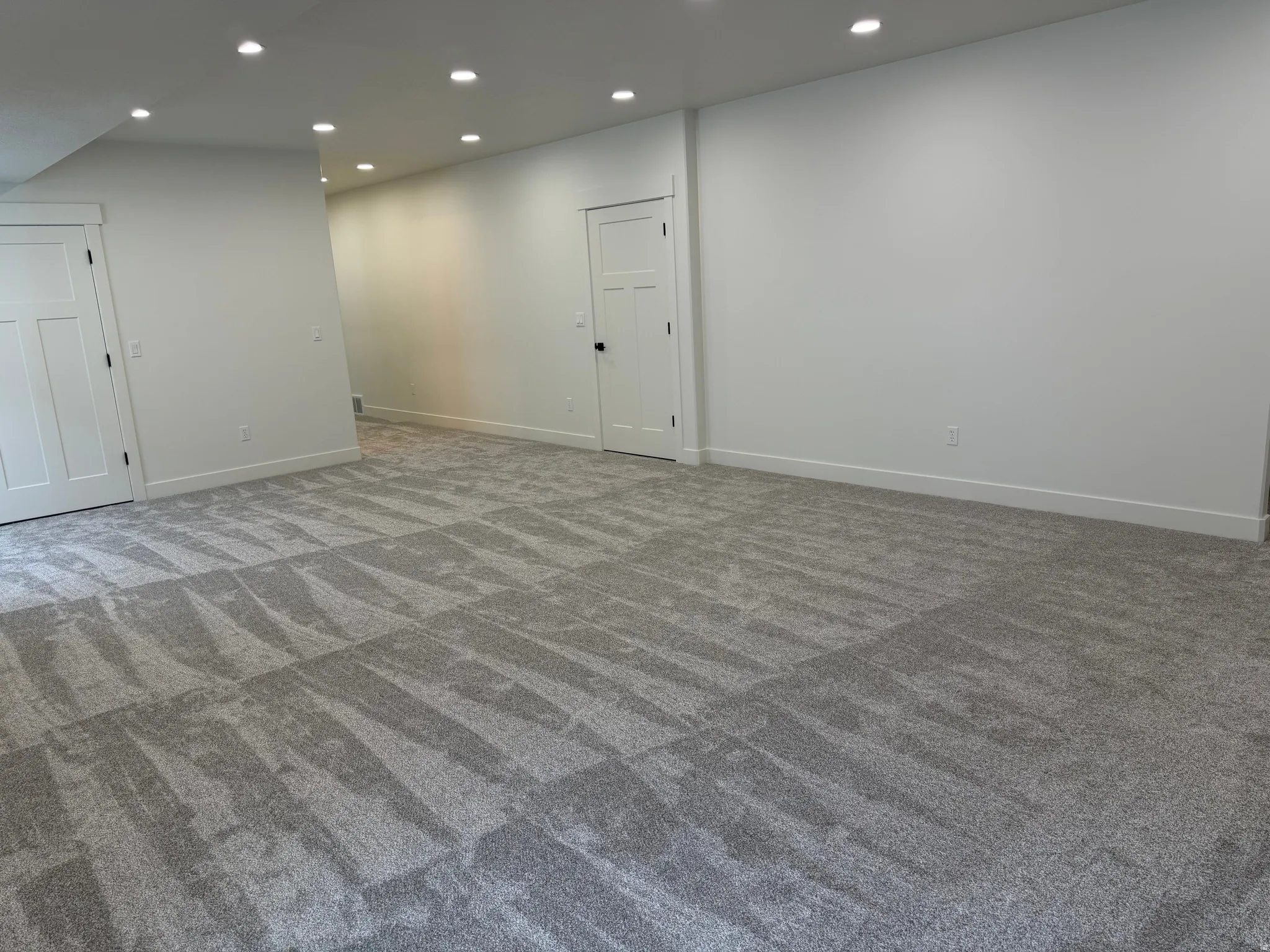 Spacious room featuring gray patterned carpet, recessed ceiling lighting, crisp white walls, and two white panel doors with black hardware