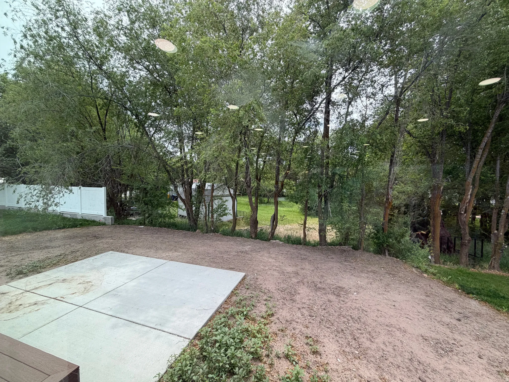 Backyard featuring a concrete patio, a sloped area ready for landscaping, a white privacy fence, and mature trees providing natural screening