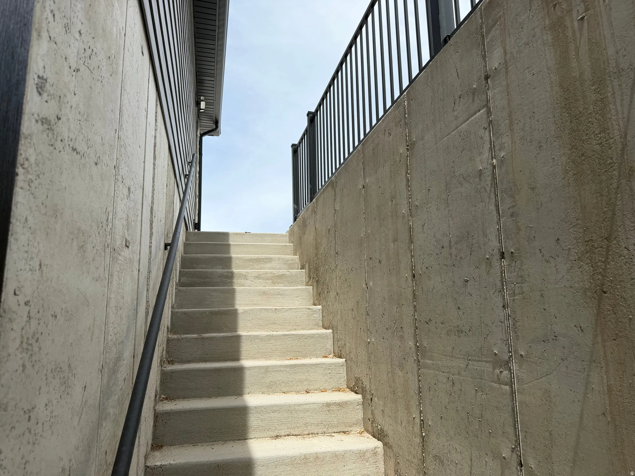 Concrete staircase with black metal railings, flanked by a concrete retaining wall and a building with dark siding and soffit