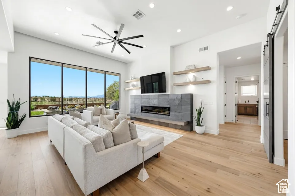 Spacious living area featuring large windows, wood-finish flooring, a linear fireplace with stone surround, a modern ceiling fan, and recessed lighting