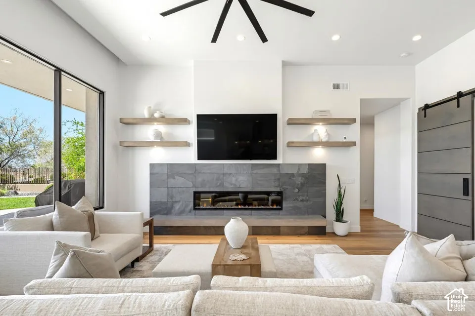 Spacious living area featuring a linear fireplace with a stone surround, wood-finish flooring, a contemporary ceiling fan, recessed lighting, and a barn door
