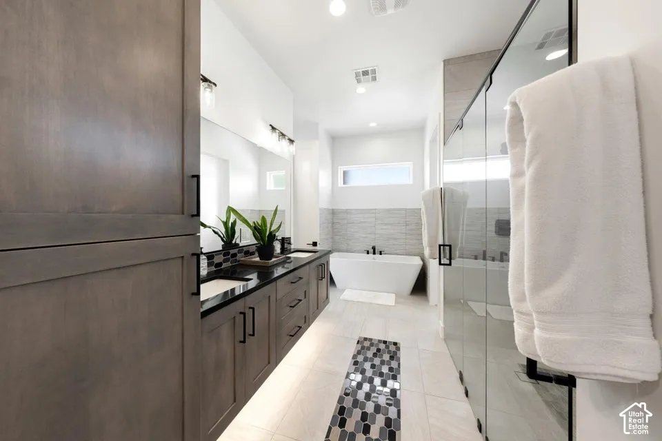 Spacious bathroom featuring a dual vanity with dark countertops and integrated sinks, a freestanding soaking tub, a separate glass-enclosed shower, and light wood-finish cabinetry