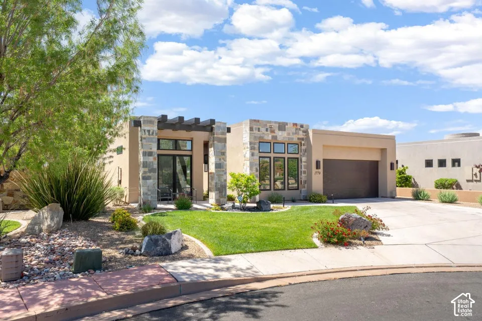 Contemporary residence featuring a stucco exterior, stone accents, an attached garage, a manicured lawn, and mature landscaping
