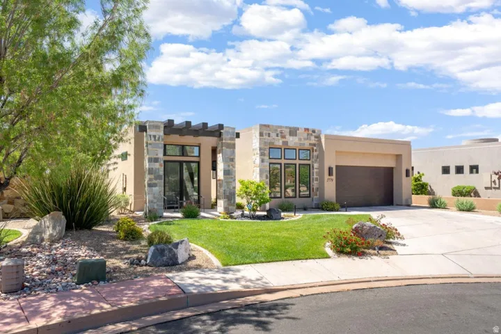 Contemporary residence featuring a stucco exterior, stone accents, an attached garage, a manicured lawn, and mature landscaping