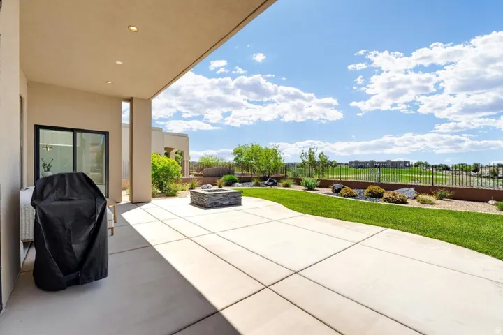 Expansive concrete patio featuring a built-in fire pit with stone-look surround, manicured lawn, and a low retaining wall with integrated landscaping