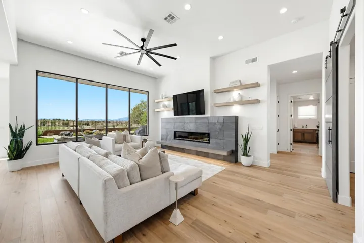 Spacious living area featuring large windows, wood-finish flooring, a linear fireplace with stone surround, a modern ceiling fan, and recessed lighting