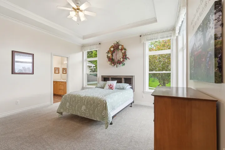 Carpeted room featuring a tray ceiling with a ceiling fan, two large windows with white trim, and a doorway opening to a bathroom with a wood-finish vanity and mirror