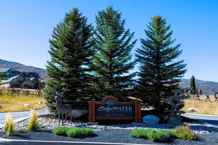 Entryway signage for "Edgewater The Chalets" featuring a wood-frame sign, three mature evergreen trees, decorative rock landscaping, and a deer statue