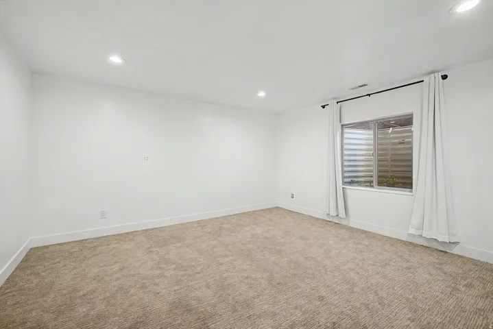 Spacious room featuring neutral carpeting, white painted walls, recessed lighting, and a window with white trim and drapes