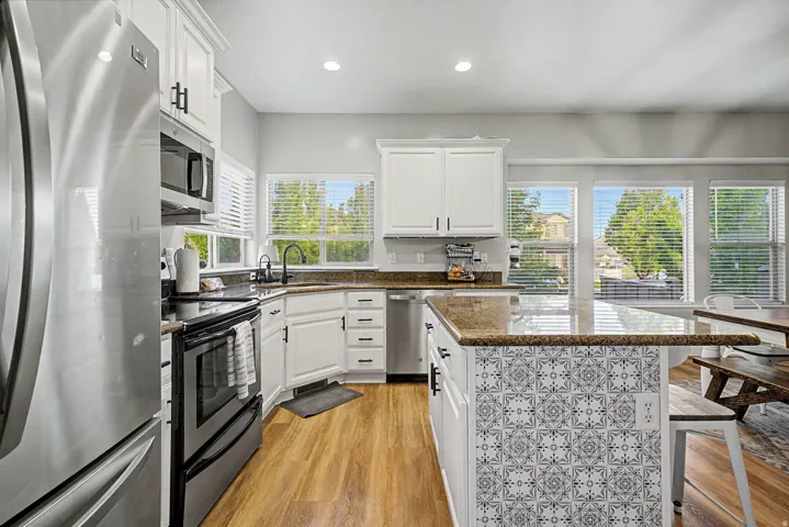 Kitchen featuring wood-finish flooring, white cabinetry with dark hardware, a stainless steel appliance suite, granite countertops, and an island with decorative tile-finish paneling