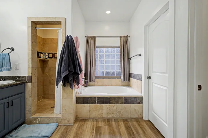 Full bathroom featuring a built-in soaking tub, a stand-up shower with tiled walls and an integrated shelf, a vanity with a dark countertop, white walls, and wood-finish flooring