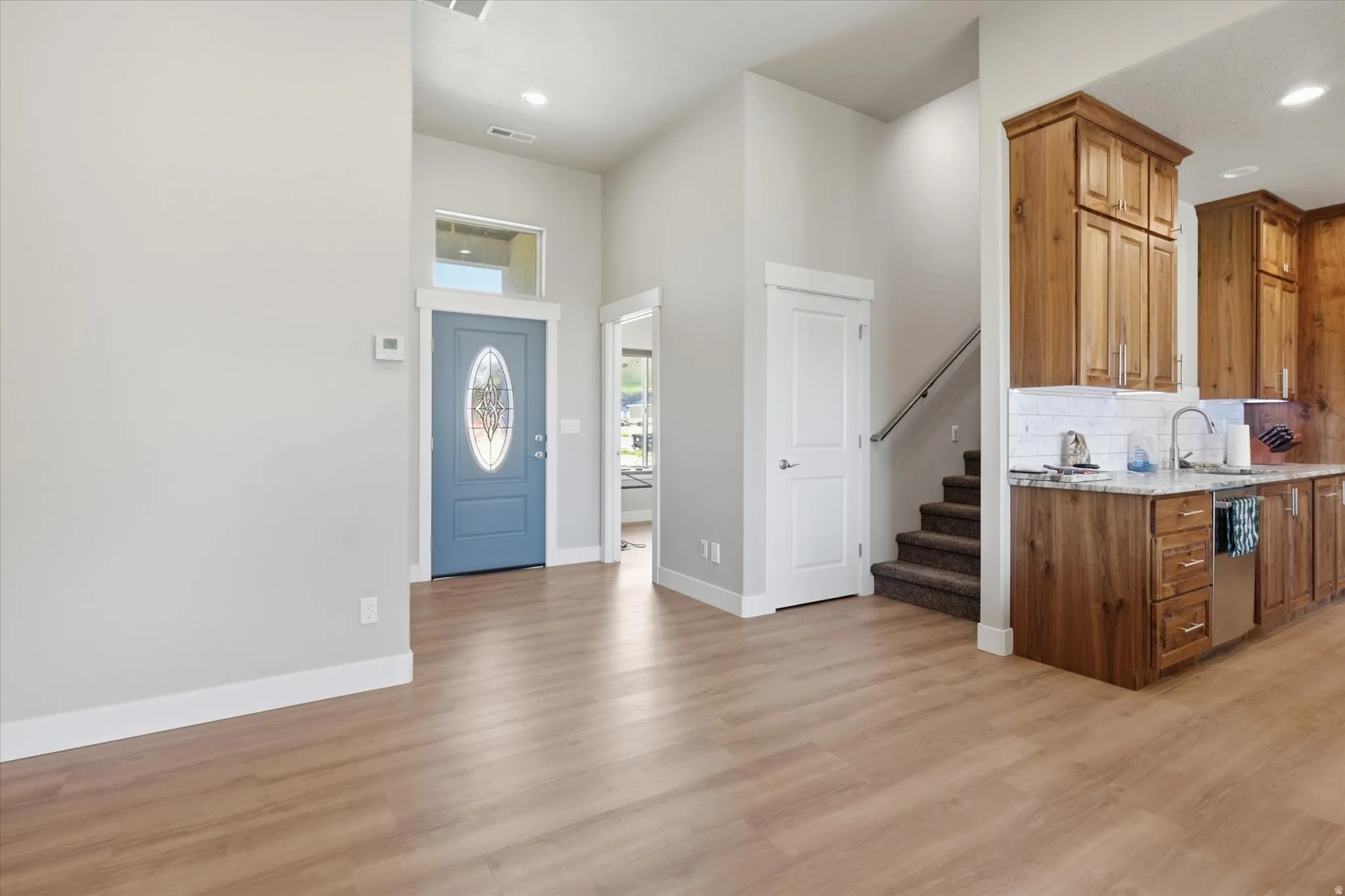 Open-concept living area with wood-finish flooring, a contemporary blue entry door with decorative glass, and a kitchen featuring wood cabinetry, light-toned countertops, and a subway tile backsplash