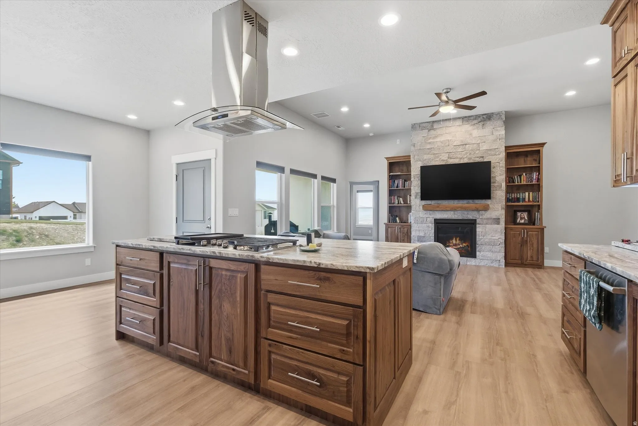 Open-concept living space featuring a large kitchen island with cooktop and stainless steel range hood, natural wood-finish cabinetry, light wood-finish flooring, and a stone-clad fireplace with built-in shelving