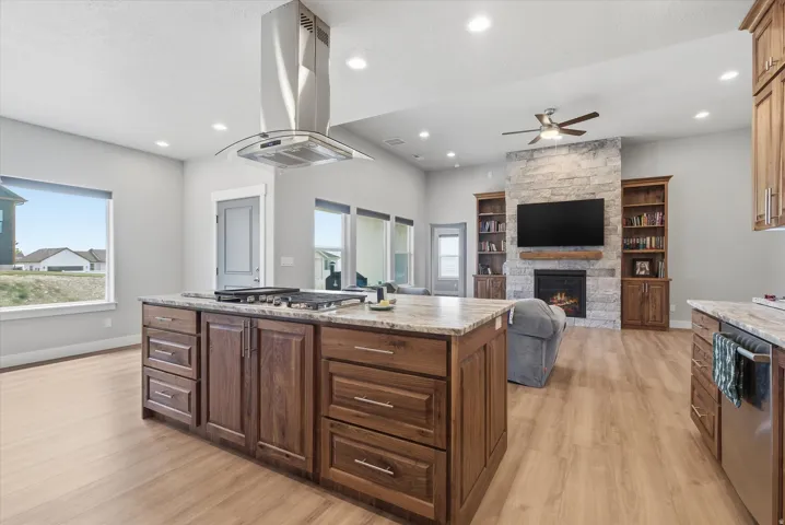 Open-concept living space featuring a large kitchen island with cooktop and stainless steel range hood, natural wood-finish cabinetry, light wood-finish flooring, and a stone-clad fireplace with built-in shelving