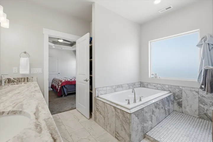 Spacious bathroom featuring a built-in tub with tile surround, a large window, tiled shower floor, and a double vanity with stone countertop