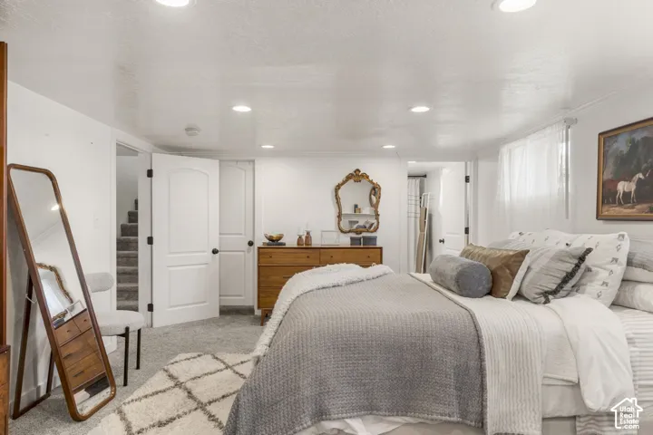 Carpeted primary bedroom featuring white walls, recessed ceiling lighting, a window with white sheer curtains, and a solid white door with black hardware