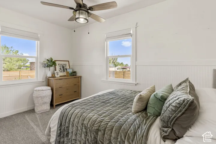Recessed lighting and a ceiling fan fixture illuminate this bedroom, featuring white wainscoting and plush carpeting