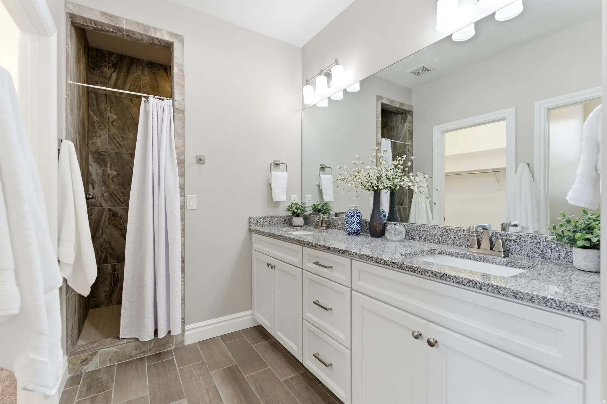 Bathroom featuring a dual vanity with granite-style countertops, wood-finish tile flooring, a walk-in shower with stone-look tile surround, and a large wall-mounted mirror