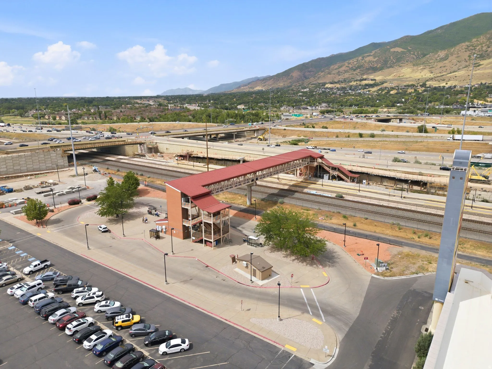 Overhead pedestrian bridge with red roofing and exterior stairs, bordered by a paved parking lot and railway lines