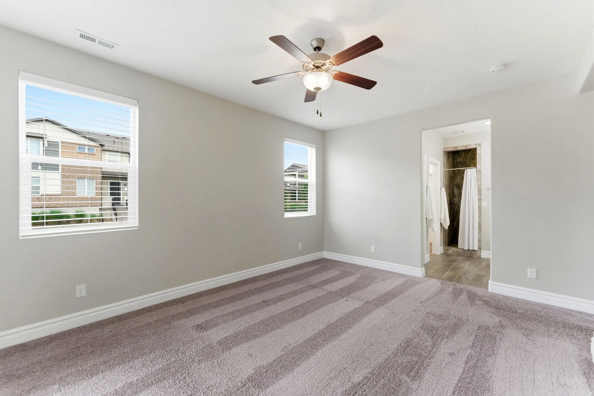 Carpeted bedroom featuring two windows with horizontal blinds, a ceiling fan with integrated lighting, and an entryway to an attached bathroom with tiled flooring and a shower stall