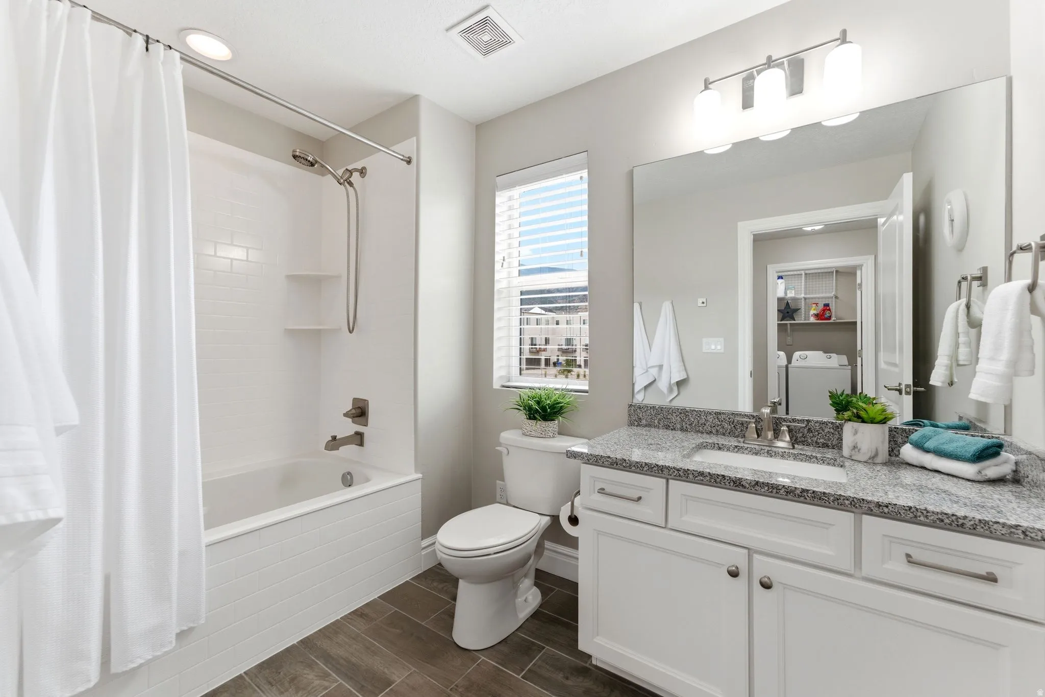 Contemporary bathroom featuring a white subway tile tub surround with integrated shelving, a single vanity with stone countertop, wood-finish flooring, a window with horizontal blinds, and a large wall-mounted mirror